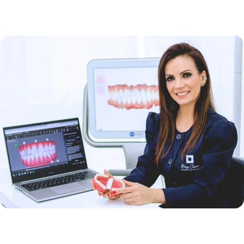 Foto de Dra Caroline sentada com um protótipo de dentes nas mãos com dois computadores ao fundo representando a simulação do seus dentes alinhados no futuro. Alinhadores Invisalign: A Solução Invisível para o Perfeito Sorriso.Clinica Geral em Vila Velha